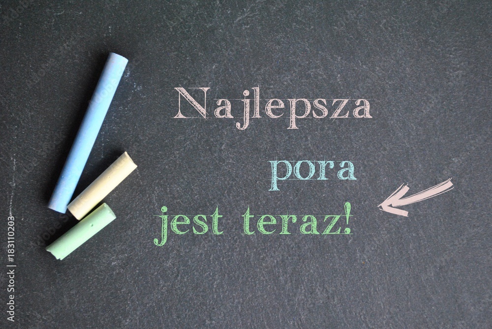 Napis 'Najlepsza pora jest teraz!' wykonany kolorową kredą na czarnej tablicy, obok leżące kolorowe kredy, symbolizujący motywację do natychmiastowego działania
