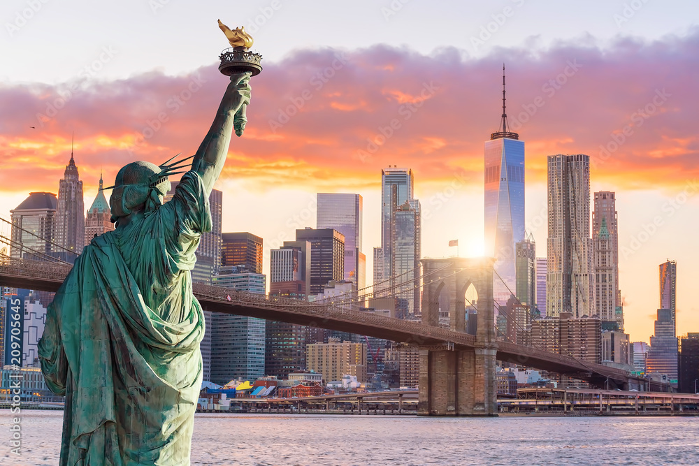 Statua Wolności na tle panoramy Nowego Jorku o zachodzie słońca. W centrum widoczny Brooklyn Bridge oraz wieżowce, w tym One World Trade Center, otoczone ciepłymi barwami nieba.