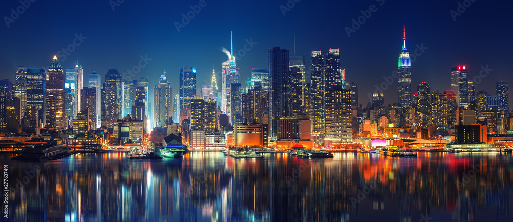 Panorama Nowego Jorku nocą z widokiem na oświetlone wieżowce Midtown Manhattan. Na pierwszym planie widać odbicia świateł w wodach rzeki Hudson, a w tle charakterystyczne Empire State Building i inne drapacze chmur.