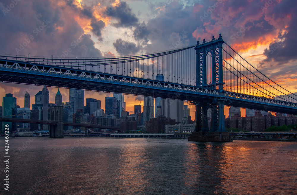 Panorama Nowego Jorku o zachodzie słońca z widokiem na Manhattan Bridge i wieżowce miasta odbijające się w wodach East River. Dynamiczne niebo pełne kolorów podkreśla kontrast między industrialną architekturą mostu a nowoczesnym stylem miejskim.