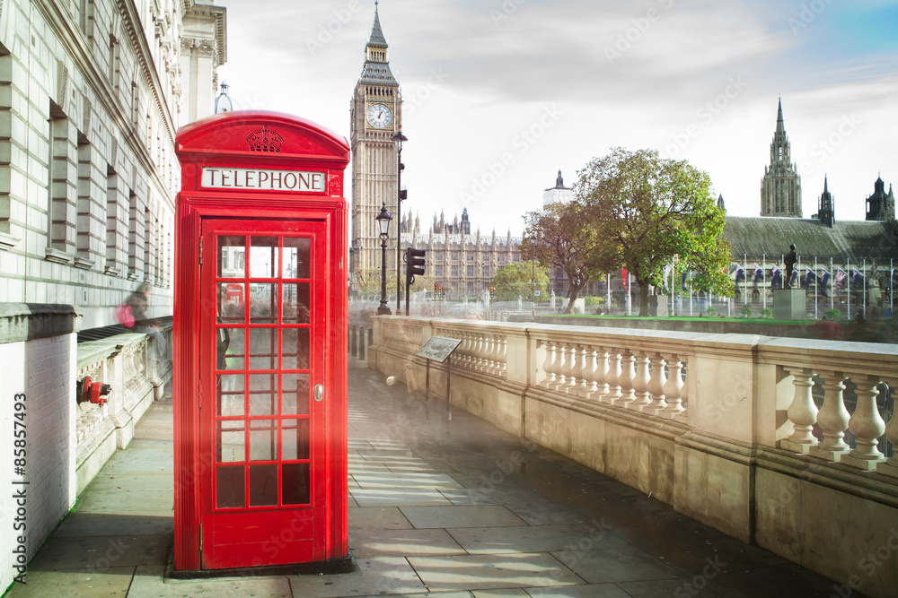Czerwona budka telefoniczna na tle Big Bena i Pałacu Westminsterskiego w Londynie. Scena ukazuje klasyczną architekturę brytyjską w słoneczny dzień, z elementami zieleni i miejskiego krajobrazu.